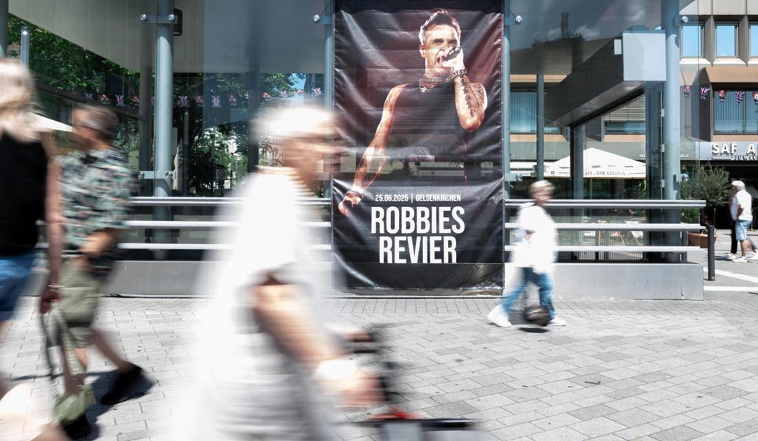 25.06.2025, Nordrhein-Westfalen, Gelsenkirchen: Ein großes Poster, auf dem „Robbies Revier“ steht, hängt in der Innenstadt. Infostände, Merchandise, Klebetattoos, Masken und Musik sollen die Fans von Robbie Williams auf das Konzert am Abend in der Veltins Arena einstimmen. Foto: Fabian Strauch/dpa - ACHTUNG: Nur zur redaktionellen Verwendung im Zusammenhang mit der aktuellen Berichterstattung und nur mit vollständiger Nennung des vorstehenden Credits +++ dpa-Bildfunk +++