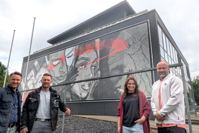 Vier Verantwortliche stehen vor einer mit einem Kunstwerk gestalteten Fassade eines Fitnessstudios.