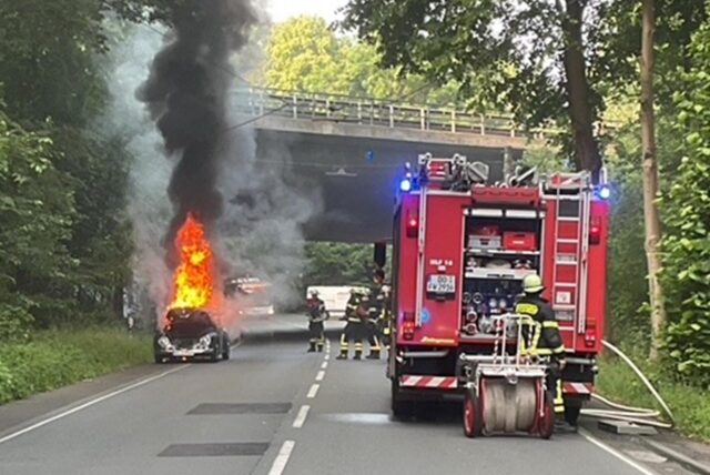 Ein VW-Käfer brennt, daneben steht ein Fahrzeug der Feuerwehr. Feuerwehrleute sind mit Löscharbeiten beschäftigt.