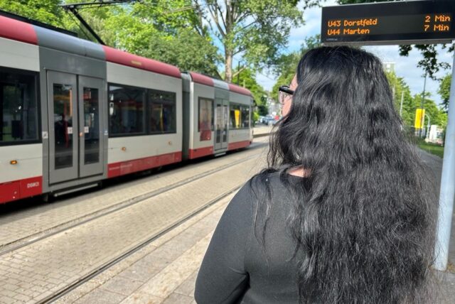 Elisa steht mit dem Rücken zur Kamera. Sie blickt auf eine Bahn die gerade an der Haltestelle Wittener Straße in Dortmund steht.
