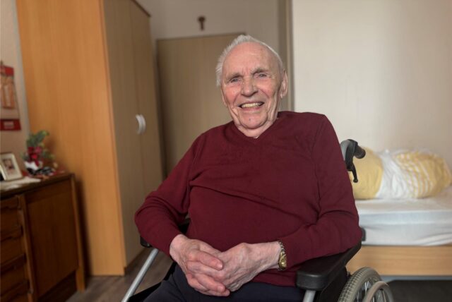 Ein älterer Mann sitzt in einem Rollstuhl in einem Zimmer. Er trägt einen roten Pullover und blickt lächelnd in die Kamera.