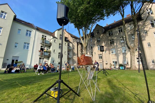 Eine Gruppe von Menschen sitzt auf einer Rasenfläche vor einem Wohngebäude. Ein Lautsprecher ist im Vordergrund zu sehen.