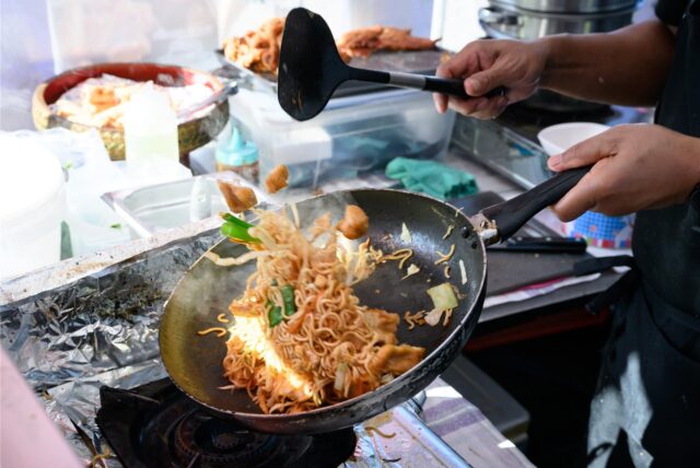 Ein Mitarbeiter eines Street-Food-Wagens schwenkt eine Pfanne mit einem Nudelgericht.