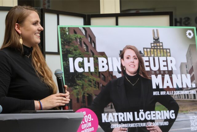 Grünen-Politikerin Katrin Lögering steht neben einem Plakat, das sie als Oberbürgermeister-Kandidatin 2025 für Dortmund zeigt.