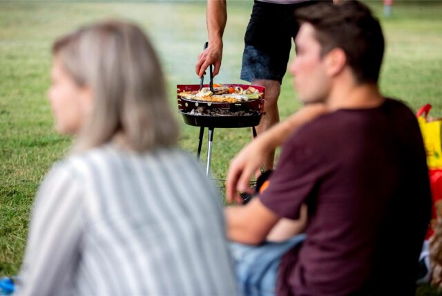 Ein Mann und eine Frau sitzen vor einem Grill auf der Wiese, während ein Dritter mit einer Zange Gegrilltes wendet.