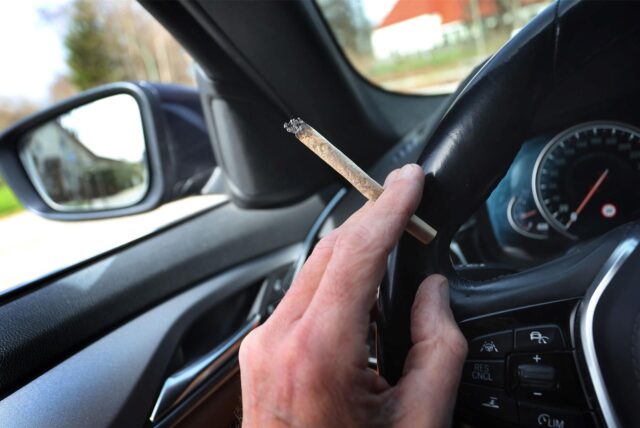 Die linke Hand ist am Steuer eines Autos. Zwischen dem Zeige- und Mittelfinger ist ein Joint. (Symbolbild)