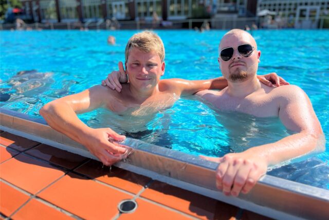 Zwei Männer im Schwimmerbecken, einer trägt eine Sonnebrille, beide sind am Beckenrand.