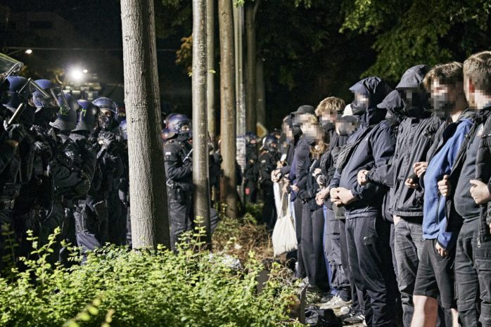 Dutzende Demonstrierende stehen einer Polizeikette gegenüber, die die Gruppe umschließt. die Demonstrierenden sind untereinander eingehakt, ihre Gesichter wurden unkenntlich gemacht.