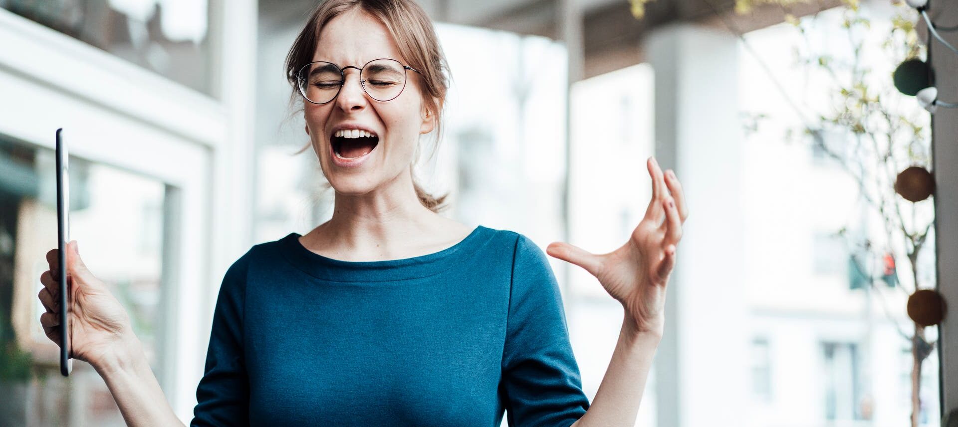 Eine junge Frau in einem blauen Oberteil steht in einem modernen Büro. Sie hält beide Arme leicht erhoben, in einer Hand ein Tablet. Ihre Mimik wirkt gequält und angestrengt: Sie hat die Augen zusammengekniffen und den Mund weit offen, als würde sie schreien.