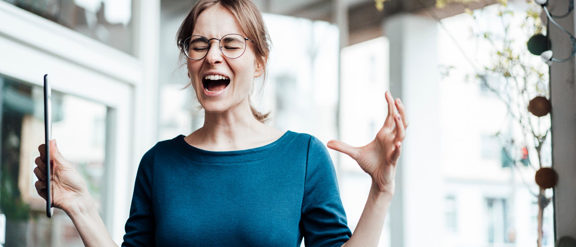Eine junge Frau in einem blauen Oberteil steht in einem modernen Büro. Sie hält beide Arme leicht erhoben, in einer Hand ein Tablet. Ihre Mimik wirkt gequält und angestrengt: Sie hat die Augen zusammengekniffen und den Mund weit offen, als würde sie schreien.