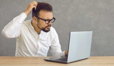 Bärtiger Mann mit Brille blickt ratlos auf Laptop und kratzt sich am Kopf.
