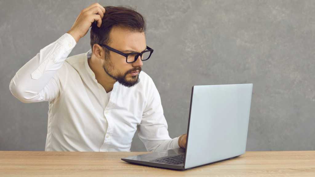 Bärtiger Mann mit Brille blickt ratlos auf Laptop und kratzt sich am Kopf.