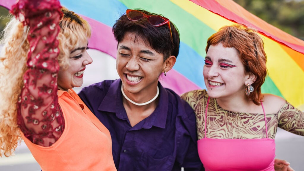 Drei fröhliche queere Menschen unter Regenbogenfahne beim CSD Köln, mit bunten Outfits und Make-up.