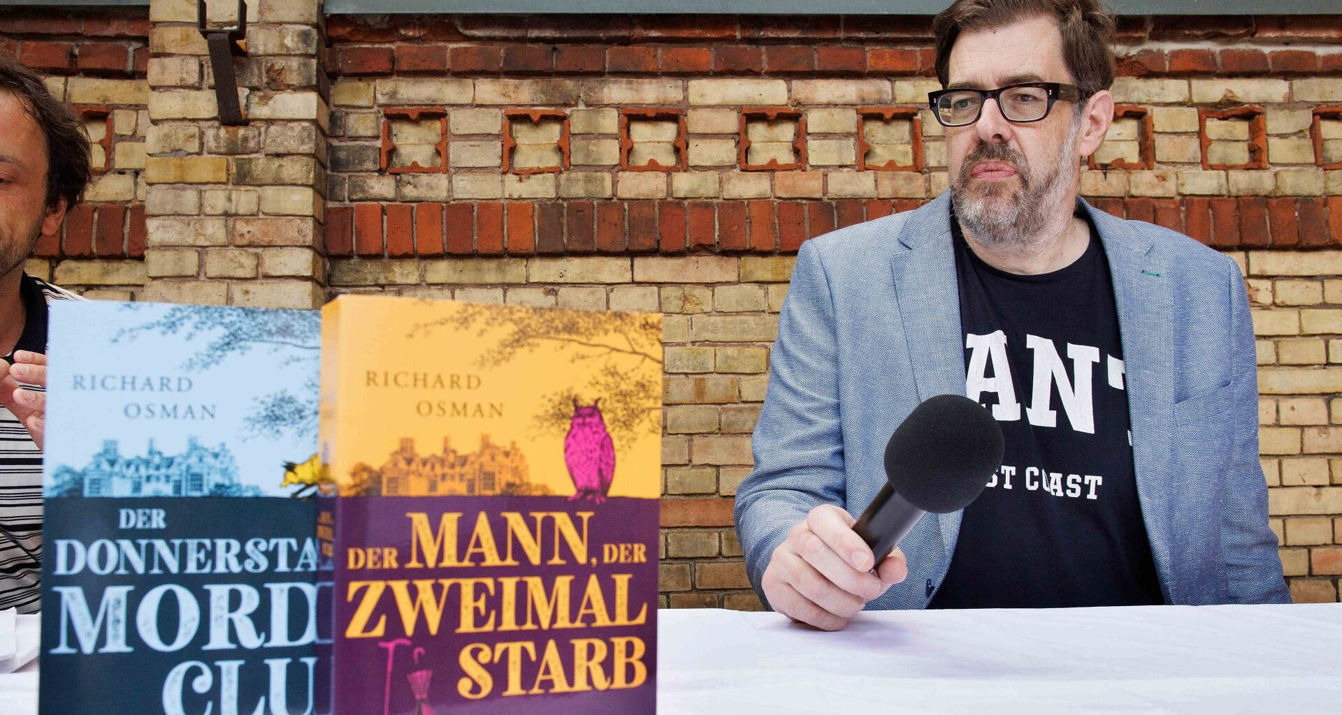 Der Bestsellerautor Richard Osman sitzt mit seinen "Der Donnerstagsmordclub"-Büchern an einem Tisch.