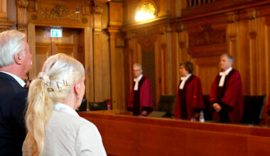 Zwei Personen, Rückenansicht im Gerichtssaal, im Hintergrund Richter in roten Roben.