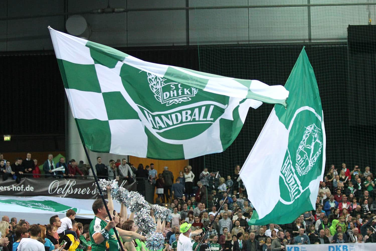 Zwei große grün-weiße Fahnen mit dem Vereinswappen des SC DHfK Handball in der Arena Leipzig. Im Hintergund sind viele Zuschauer zu sehen. Foto: Jan Kaefer