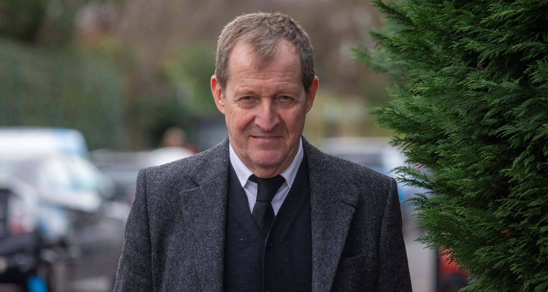 Alastair Campbell vor der St. Mary The Virgin Church in London. Er trägt einen grauen Anzug, einen dunklen Cardigan, ein weißes Hemd und eine schwarze Krawatte. Im Hintergrund sind Büsche und Autos zu erkennen.