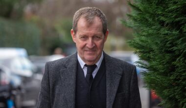 Alastair Campbell vor der St. Mary The Virgin Church in London. Er trägt einen grauen Anzug, einen dunklen Cardigan, ein weißes Hemd und eine schwarze Krawatte. Im Hintergrund sind Büsche und Autos zu erkennen.