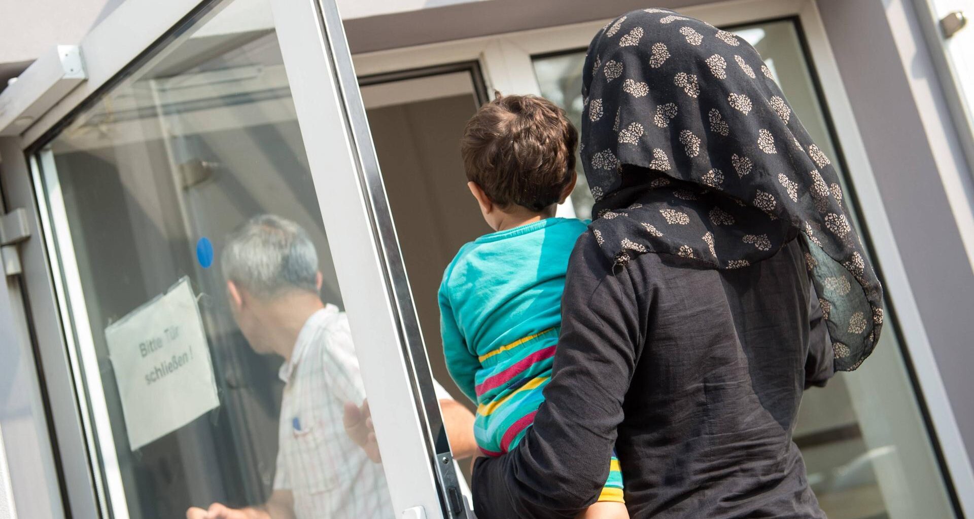 Eine verschleierte Frau mit einem Kleinkind auf dem linken Arm geht im August 2015 in Richtung einer geöffneten Tür, auf der ein Schild steht "Bitte Tür schließen". Die Tür gehört zur Ausländerbehoerde und BAMF-Außenstelle in Eisenhüttenstadt.