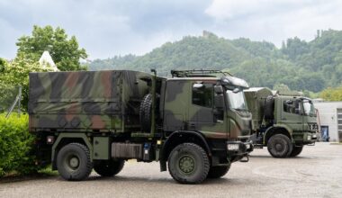 Die beiden in Tarnfarben lackierten Lkw stehen nebeneinander auf einem Parkplatz. Im Hintergrund Wald und Hecken.
