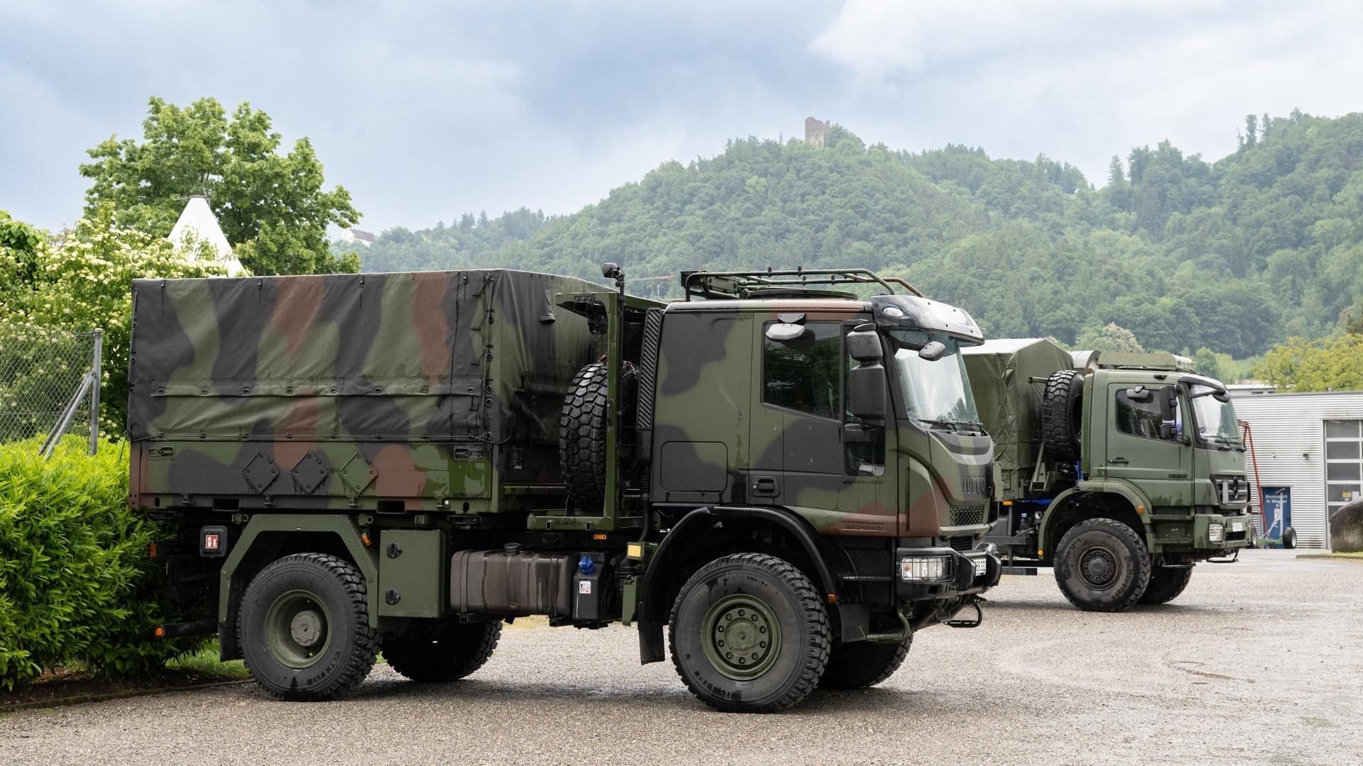 Die beiden in Tarnfarben lackierten Lkw stehen nebeneinander auf einem Parkplatz. Im Hintergrund Wald und Hecken.