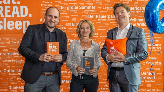 Die drei Hosts vom NDR Bücherpodcast eat.READ.sleep. Daniel Kaiser, Katharina Mahrenholtz und Jan Ehlert vor einer orangefarbenen Wand mit vielen weißen Wörtern - beim Fanfest zum 5. Geburtstag © NDR / Axel Herzig Foto: Axel Herzig