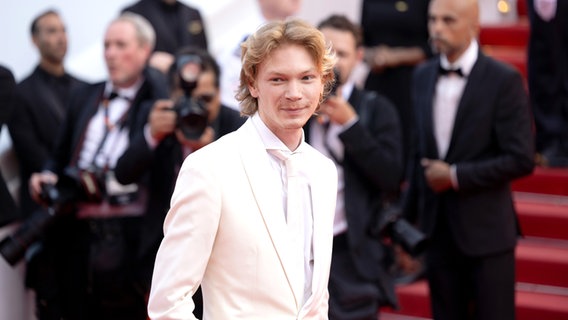 Ein junger Mann mit blonden Haaren im weißen Anzug mit weißer Krawatte  - Filip Schnack bei der Abschlussgala zum Filmfest Cannes © picture alliance / PIC ONE / Christian Ender Foto: Christian Ender