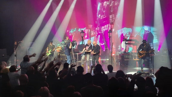 Ein Junge sitzt mit seiner Mutter auf einem Sofa auf der Bühne neben Musikern der Band Fury In The Slaughterhouse. Die Band spielt live, einer der Sänger steht am Mikrofon, ein Gitarrist spielt, im Hintergrund sitzt ein Keyboarder. Die Bühne ist in blau-violettes Licht getaucht. © NDR Foto: Uli Kniep
