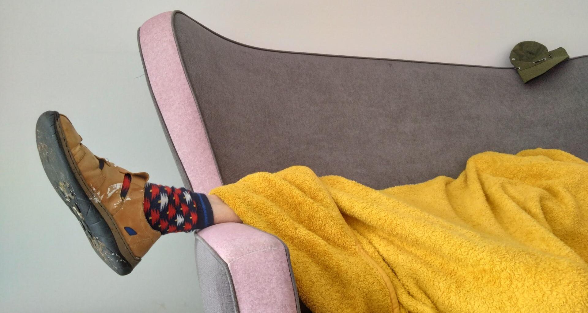 Eine Person liegt zugedeckt auf einem Sofa. Unter der Decke schaut ein Fuß heraus, der in einem Hausschuh und bunt gemusterten Socken steckt.