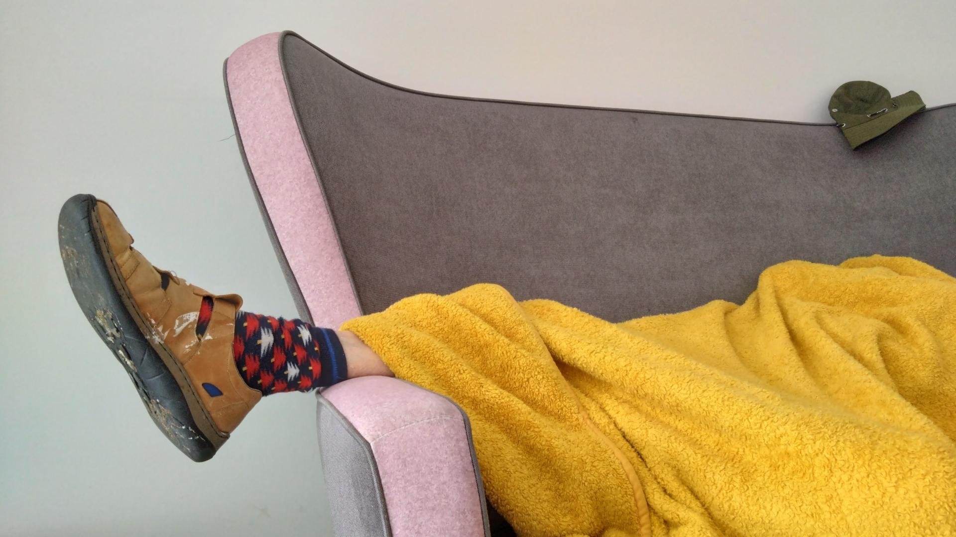 Eine Person liegt zugedeckt auf einem Sofa. Unter der Decke schaut ein Fuß heraus, der in einem Hausschuh und bunt gemusterten Socken steckt.