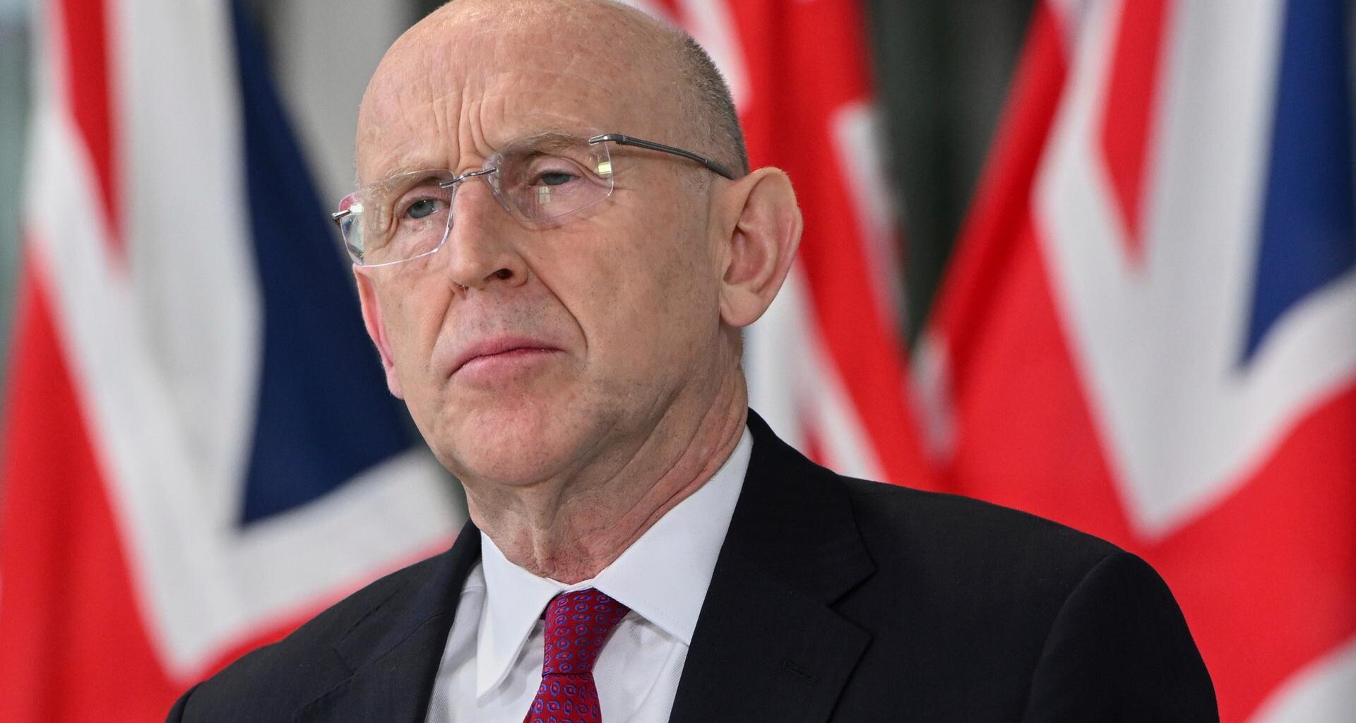 Der britische Verteidigungsminister John Healey in Anzug und Krawatte. Er trägt eine Brille. Im Hintergrund ist die britische Fahne zu sehen.