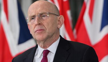 Der britische Verteidigungsminister John Healey in Anzug und Krawatte. Er trägt eine Brille. Im Hintergrund ist die britische Fahne zu sehen.