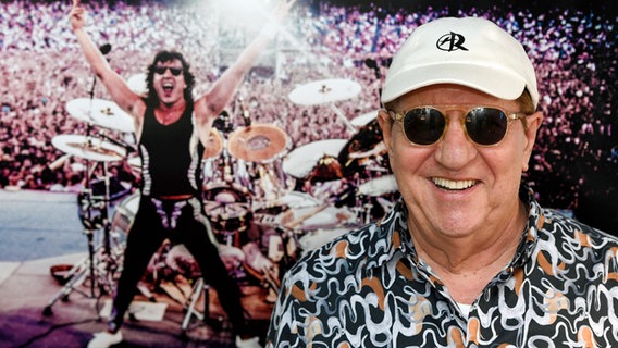 Ex-Scorpions-Drummer Herman Rarebell steht lachend vor einem großformatigen Foto aus seiner aktiven Zeit auf der Bühne. Im Hintergrund sieht man ihn am Schlagzeug, Arme in die Höhe gerissen, vor einem riesigen Konzertpublikum. © picture alliance / Geisler-Fotopress | Clemens Niehaus/Geisler-Fotopres 