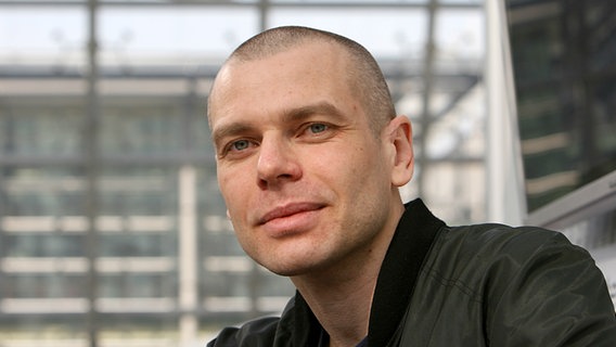 Ein Mann mit fast kahlrasiertem Kopf schaut in die Kamera. Hinter ihm ist ein Gebäude mit Glasfassade zu sehen. Es ist der Autor Wolfgang Herrndorf auf einem Bild aus dem Jahr 2007. © dpa - picture alliance Foto: Erwin Elsner