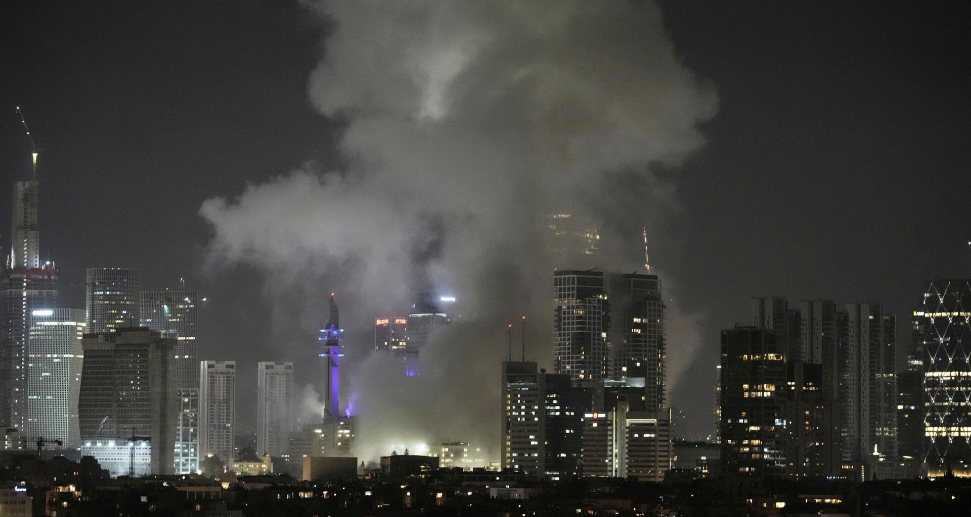 Es ist Nacht. Man sieht Gebäude und die Lichter Tel Avivs aus der Ferne. Dichter grauer Rauch steigt über Häusern auf.