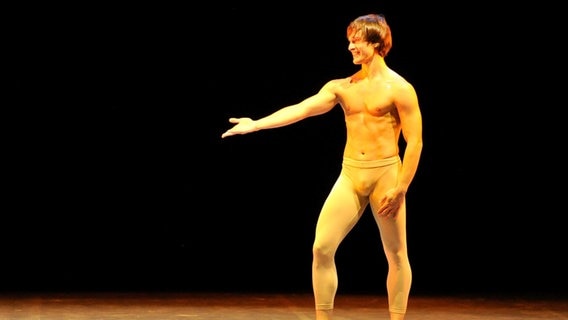 Ein Mann steht oberkörperfrei auf einer Bühne, er trägt eine helle, enge Leggins und streckt den rechten Arm nach rechts unten, dabei lacht er. © picture alliance / NurPhoto | Lorenzo Di Cola Foto: Lorenzo Di Cola