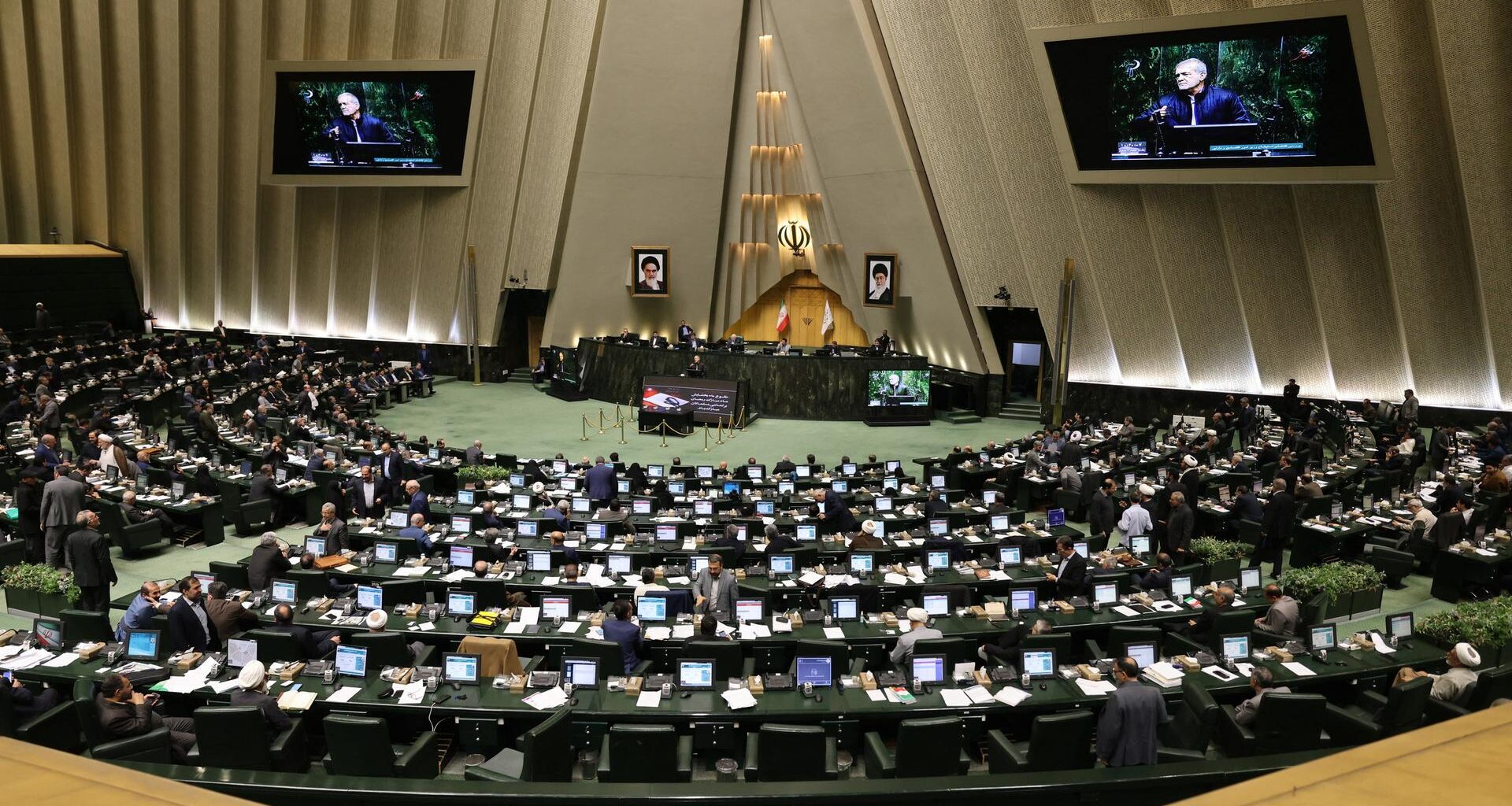 Irans Präsident Masud Peseschkian spricht zu den Abgeordneten im halbrunden Plenum. Seine Rede wird auf zwei Bildschirmen gezeigt.