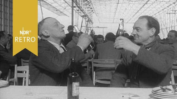 Zwei Festgäste beim Richtfest in einem Hangar prosten sich an Biertischen zu (1954). © Screenshot 