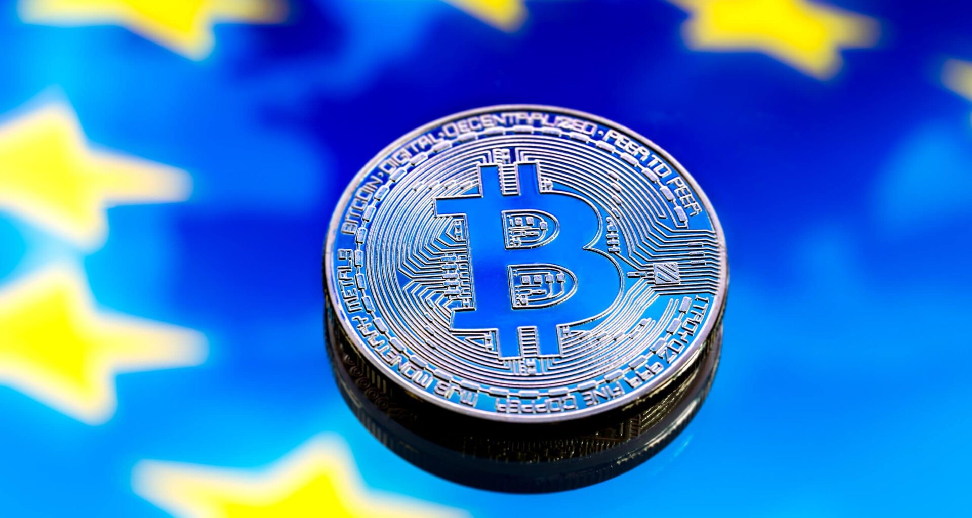 Eine physische Bitcoin-Münze liegt auf einem blauen Hintergrund mit gelben Sternen, die der Flagge der Europäischen Union ähneln und die wachsende Verbindung zwischen Krypto und der europäischen Finanzlandschaft symbolisieren.