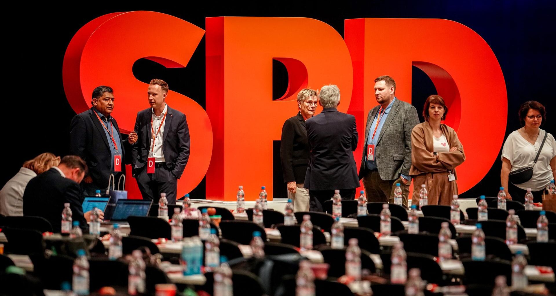 Auf der Bühne vor einem SPD-Schriftzug aus stehenden großen roten Buchstaben stehen mehrere Teilnehmer und reden. Im Vordergrund unscharf die Delegiertenbänke.