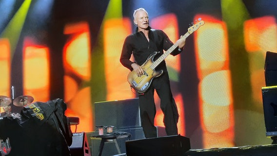 Sting spielt bei seinem Konzert in Hannover Bassgitarre, ganz in Schwarz gekleidet, vor einer farbig leuchtenden Bühnenwand mit gelb-roten Lichteffekten. © NDR Foto: Uli Kniep
