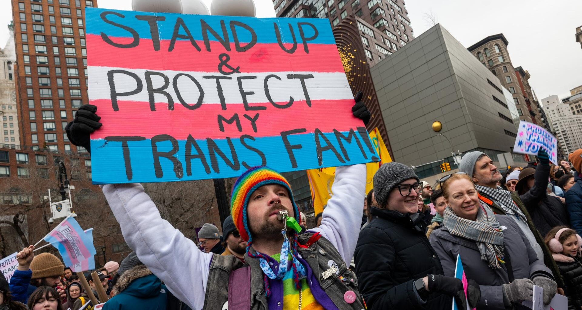 Eine Person mit vielen Regenbogenaccessoires an der Kleidung, einer Sonnenbrille und einem Bart, hält ein Schild hoch, auf dem vor den Farben der Transgender-Pride-Fahne steht „Stand Up & Protect my Trans Fam!“.