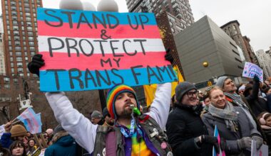 Eine Person mit vielen Regenbogenaccessoires an der Kleidung, einer Sonnenbrille und einem Bart, hält ein Schild hoch, auf dem vor den Farben der Transgender-Pride-Fahne steht „Stand Up & Protect my Trans Fam!“.