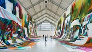 Blick in eine hohe Ausstellungshalle der Deichtorhallen Hamburg. Links und rechts hängen riesige, bunt besprühte Stoffbahnen, die vom Dach bis auf den Boden reichen. Mehrere Menschen gehen durch den Raum. © VG Bildkunst-Bonn, 2025 Foto: Henning Rogge