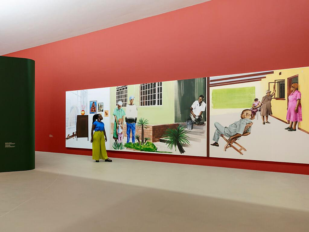 Eine Person betrachtet grosse Gemälde in einer Kunstgalerie mit roten Wänden, die Alltagsszenen darstellen.