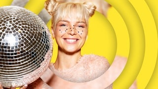 Eine blonde junge Frau mit kleinen Sternen im Gesicht hält eine Disokugel in der Hand.