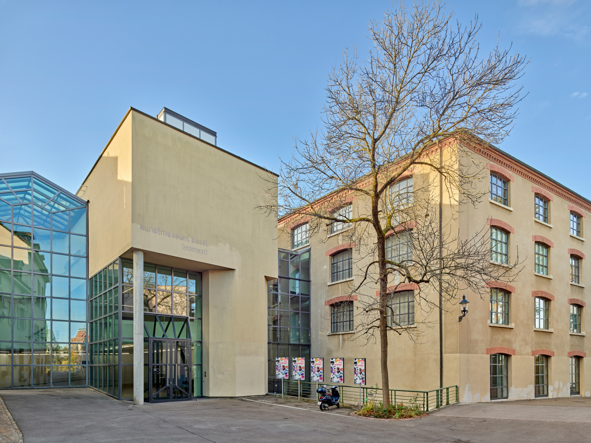 Modernes Gebäude mit Glasfassade neben einem älteren, renovierten Gebäude mit grossen Fenstern. Ein Baum steht dazwischen.
