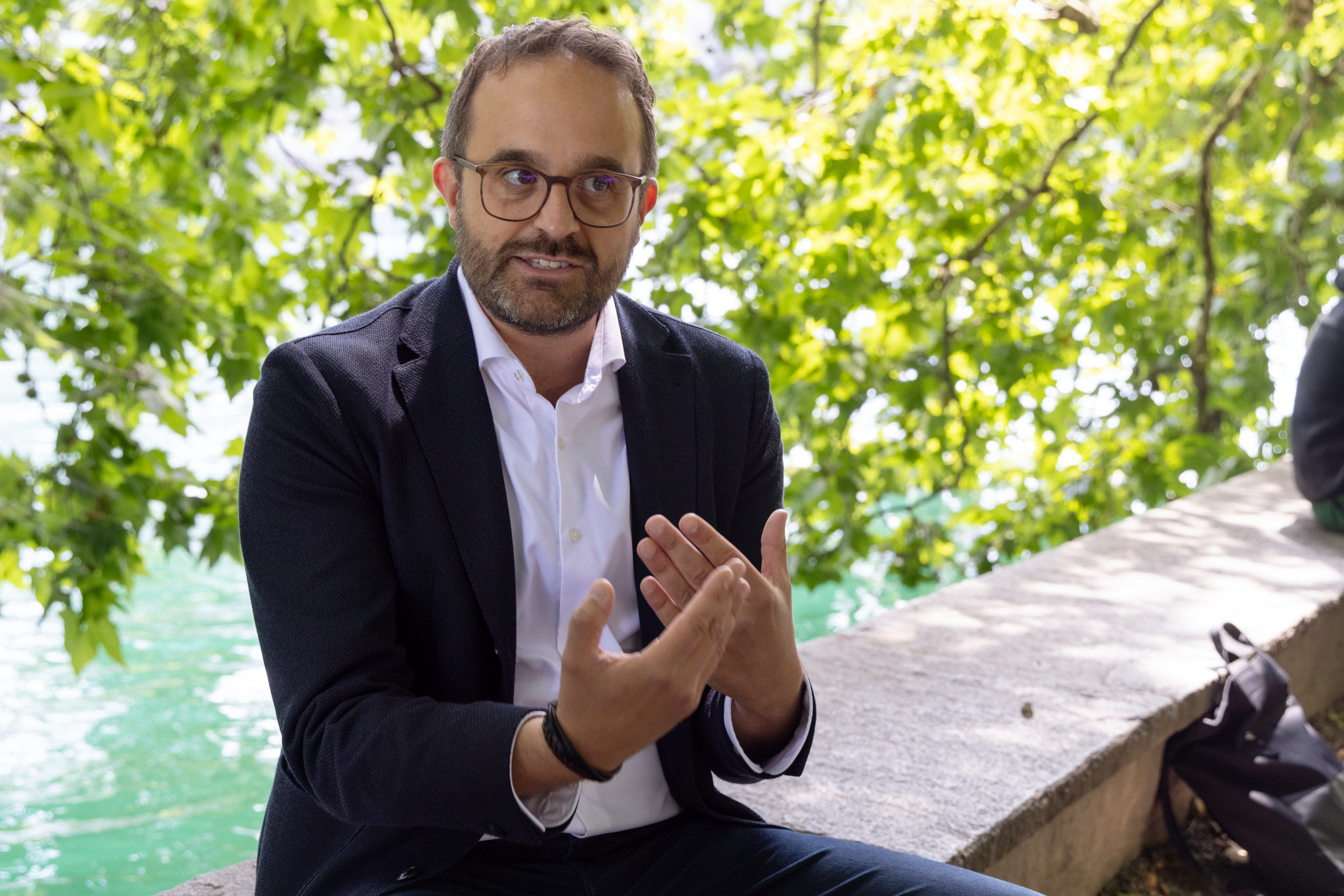 Lukas Geiger sitzt auf einer Mauer, gestikuliert beim Sprechen, im Hintergrund grüne Blätter und ein Fluss. Thema: Leben mit ADHS.