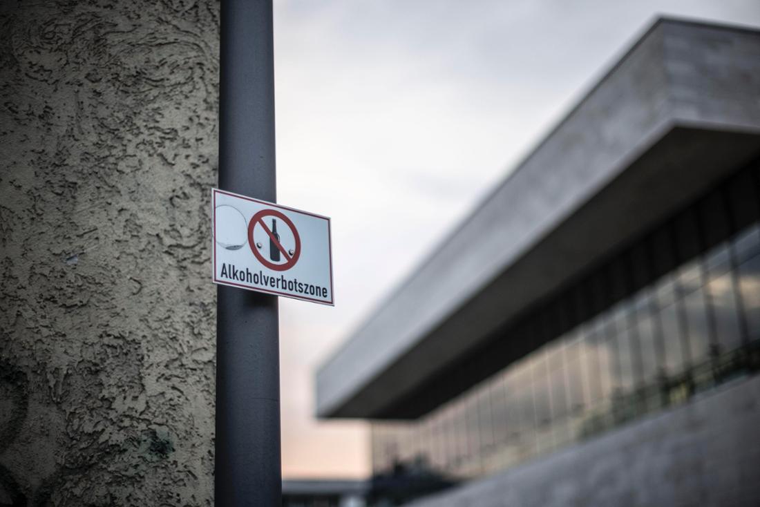 Im Vordergrund weist ein Schild an einem Mast auf die Alkoholverbotszone hin. Im Hintergrund ist die Sporthalle am Platz der Deutschen Einheit in Wiesbaden zu sehen. 