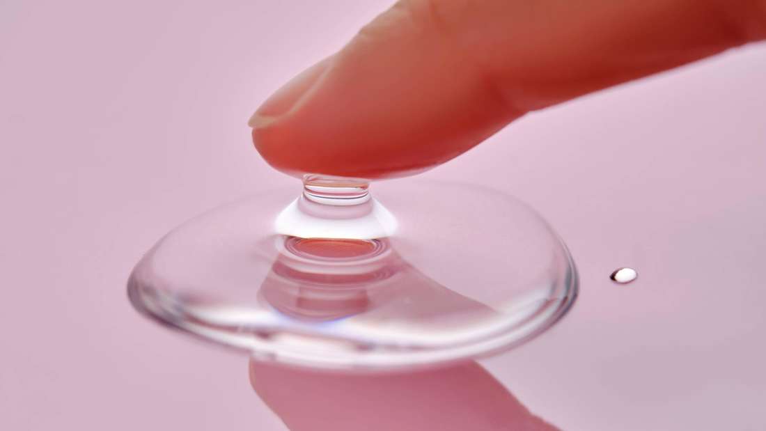 A finger touches a drop of cosmetic product on a pink background. 